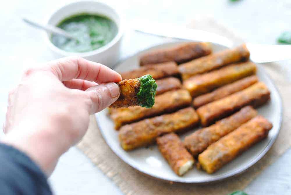 a hand holding a paneer finger dipped in green chutney over a plate of paneer fingers 