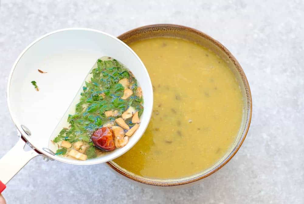 A bowl of dal with a frypan with oil, garlic, cilantro and red chilli poised over it