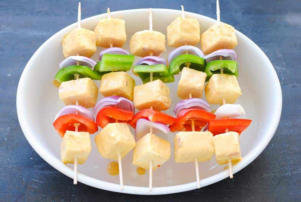 paneer, red pepper, green pepper and onion cubes on skewers resting on a white bowl