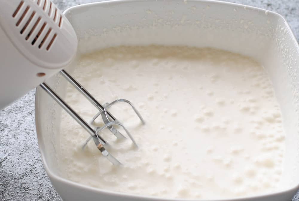 cream in a square white bowl being whipped with hand held beaters
