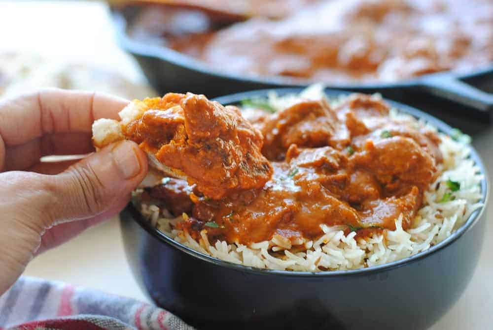 a hand scooping some chicken tikka masala from a bowl with a piece of naan