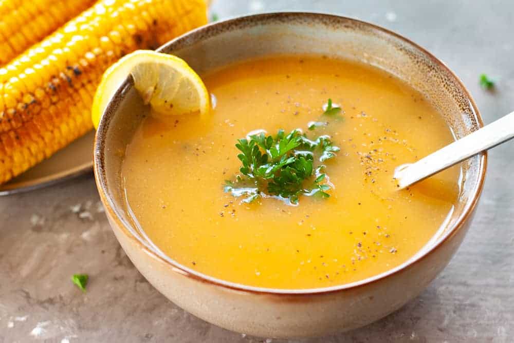 chicken and veggie soup in a bowl with corn on the cob near it