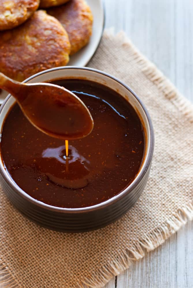 tamarind chutney in bowl with spoon over it. Fried potato patties in the background