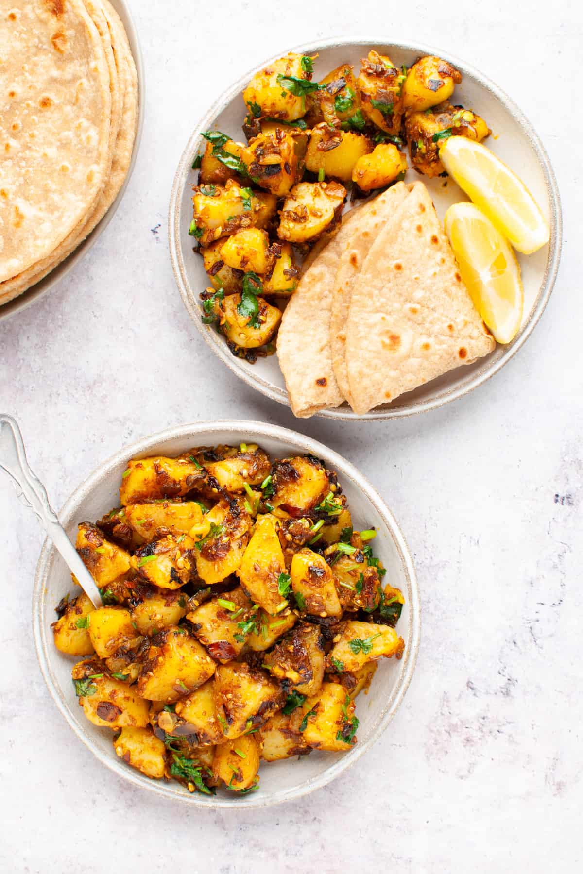 A plate with aloo sabzi and spoon; another plate with aloo sabzi, roti, and lemon wedges.