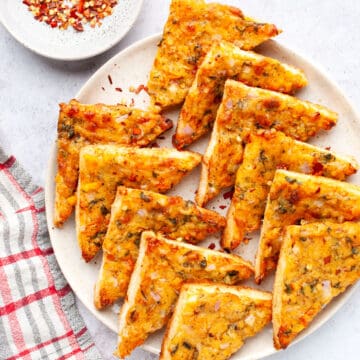 Chilli cheese toast triangles on a platter with chilli flakes on top left.